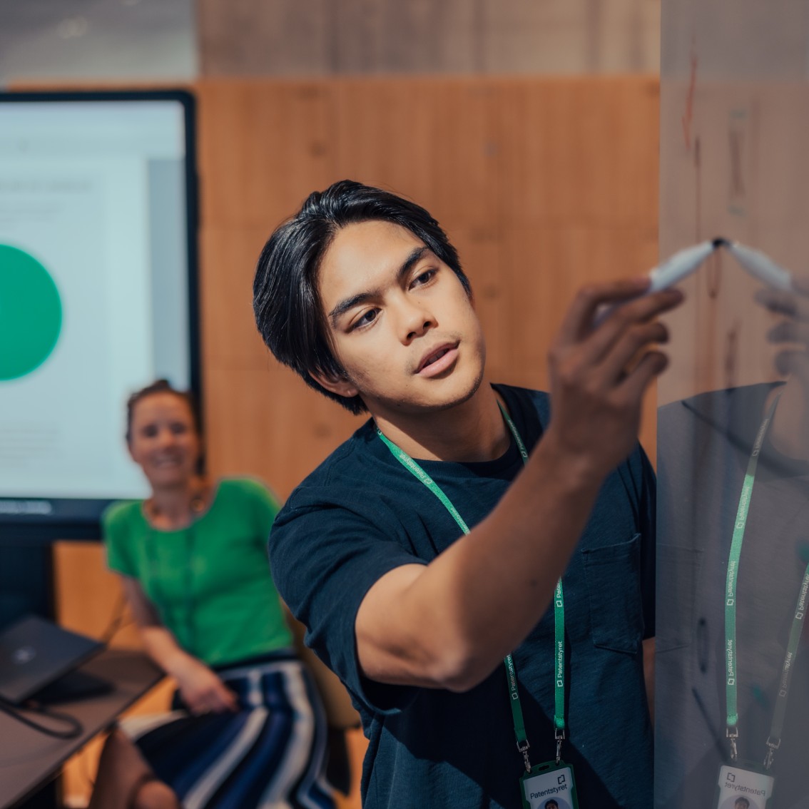 Patentstyrets ansatte samarbeider ved en whiteboard, der en person skriver med tusj mens en kollega følger med fra sidelinjen. I bakgrunnen vises en skjerm med et fargerikt sektordiagram.