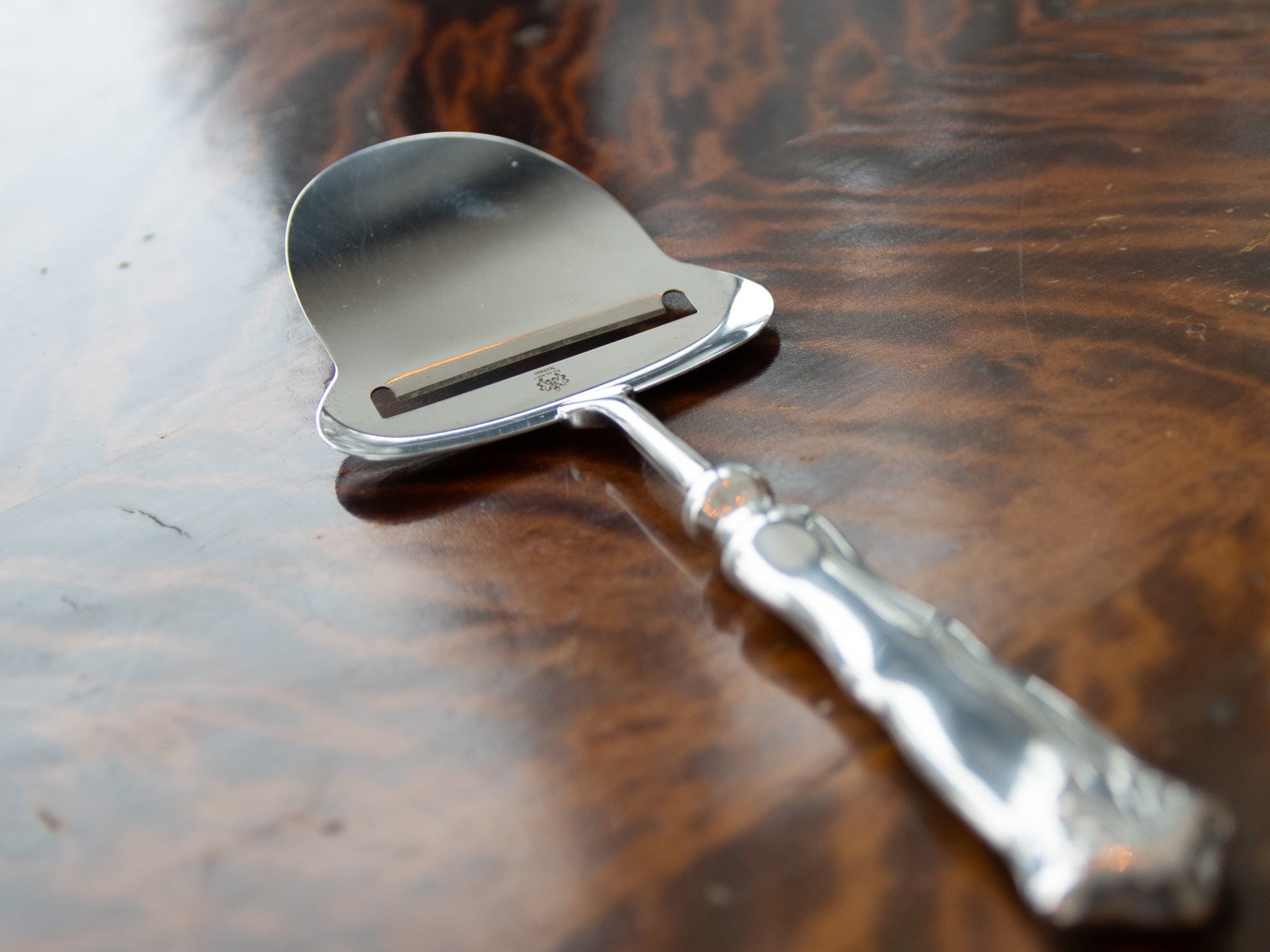 Cheese slicer for brown cheese in stainless steel, designed by Thor Bjørklund, lying on a dark wooden table.
