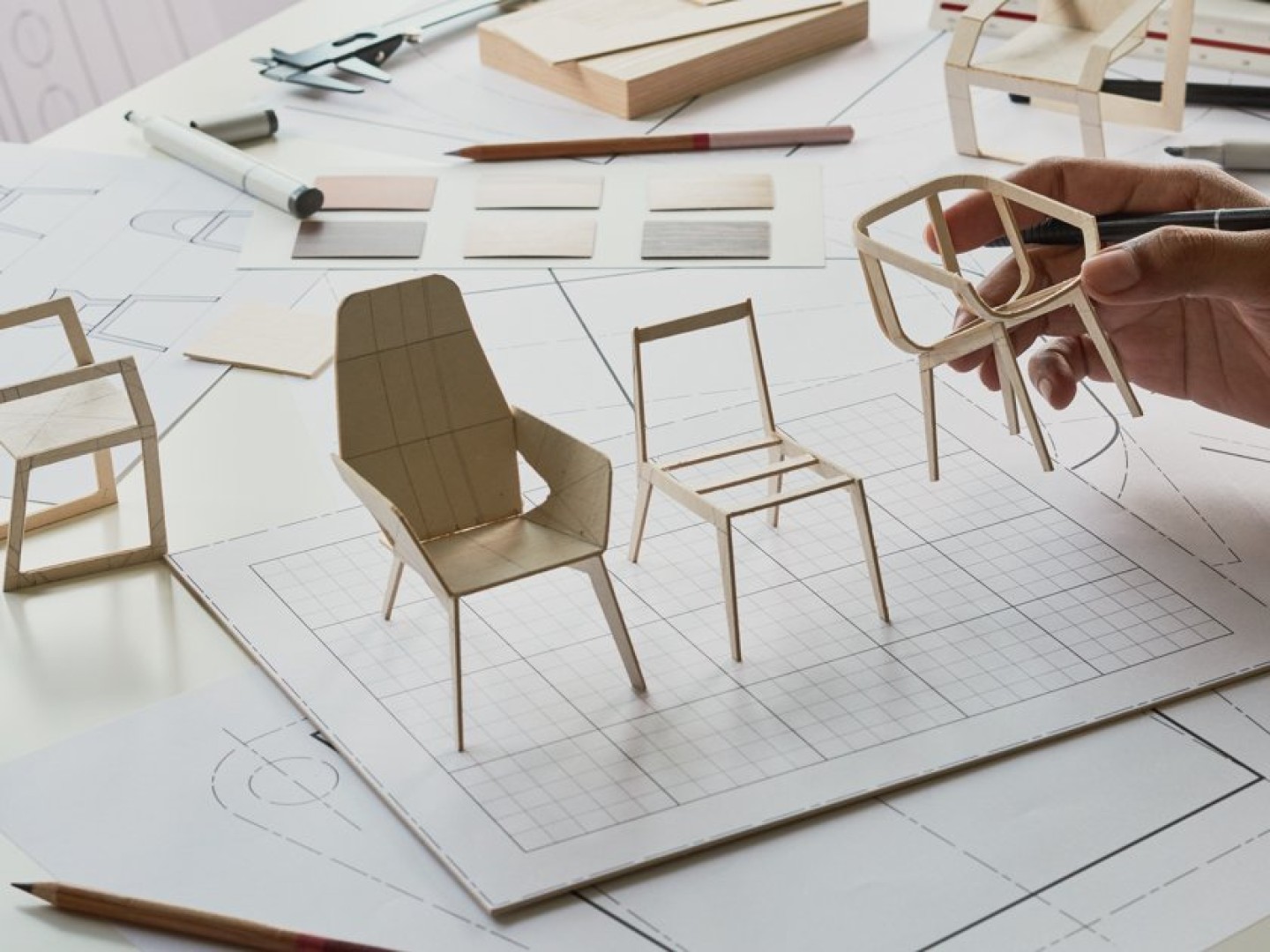 Hand holding chair model over blueprints and prototypes on a design desk, showing a design process.