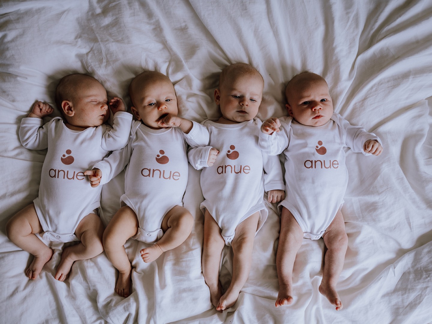 Four babies in a bed with bodysuits marked with Anue.
